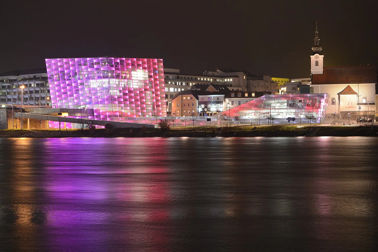 The Ars Electronica Center lit up in purple across the Danube in Linz at night.